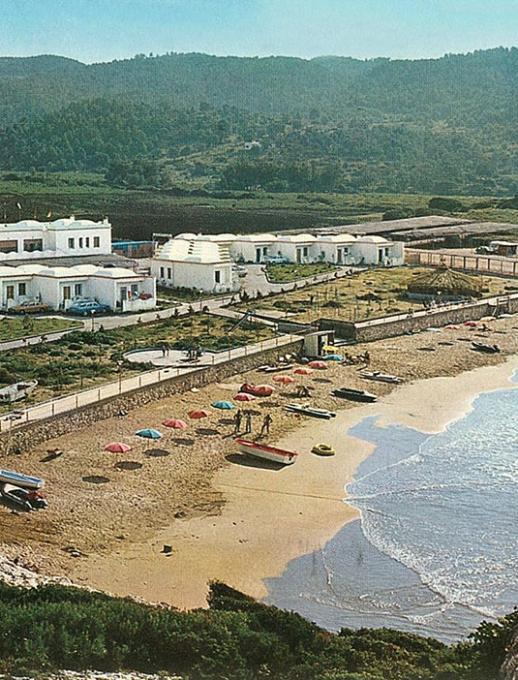 Spiaggia con ombrelloni colorati, mare mosso e complesso di edifici bianchi sullo sfondo.