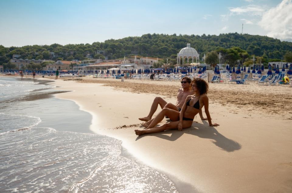 Spiaggia affollata con ombrelloni, mare cristallino e verdeggiante paesaggio costiero.